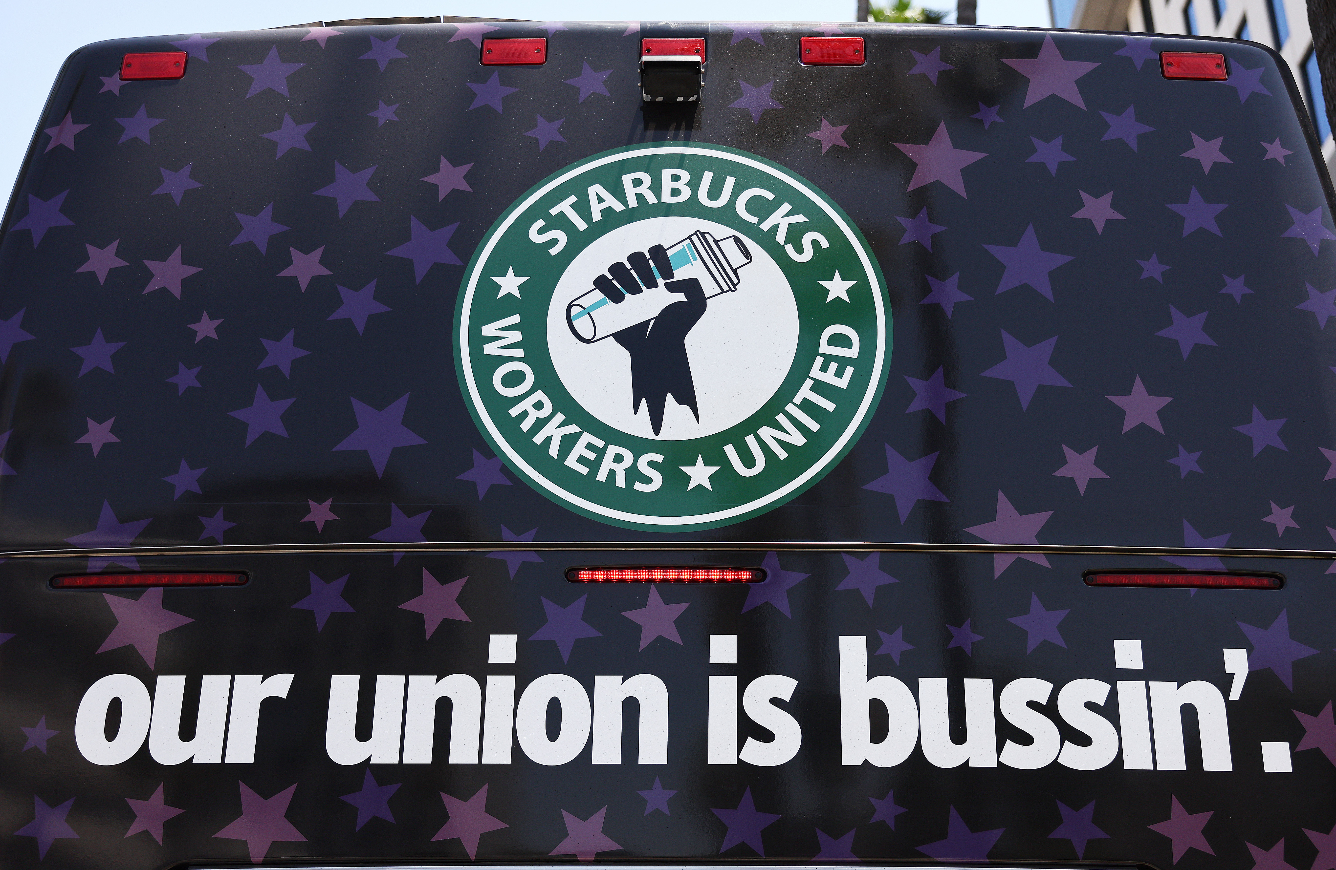 A logo featuring a hand holding a cup surrounded by the words “Starbucks Workers United.” Below the logo there are words reading “Our union is bussin’.”