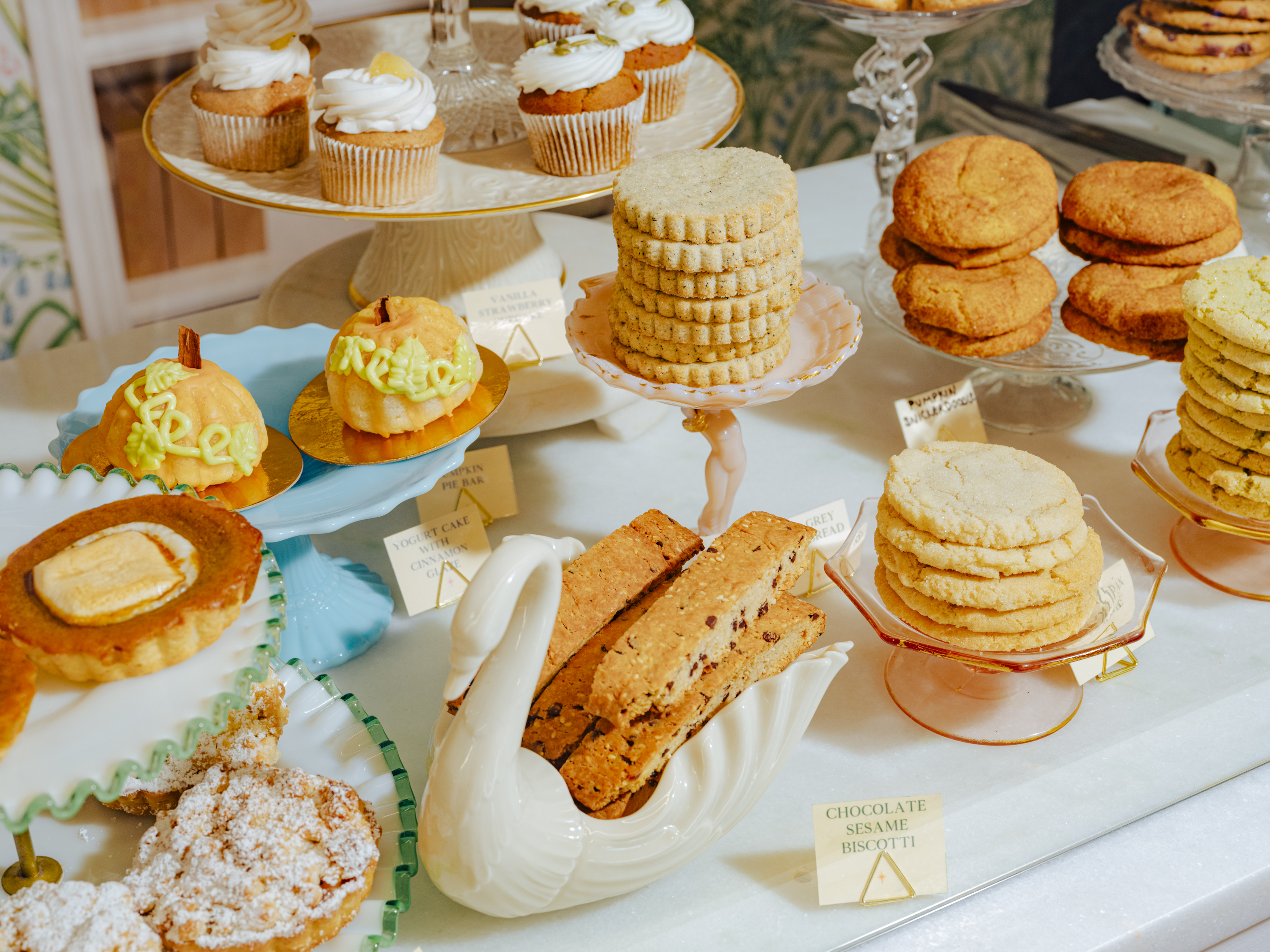 A counter filled with vegan pastries, including a ceramic swan filled with biscotti and stacks of cookies and cupcakes displayed on cake stands.