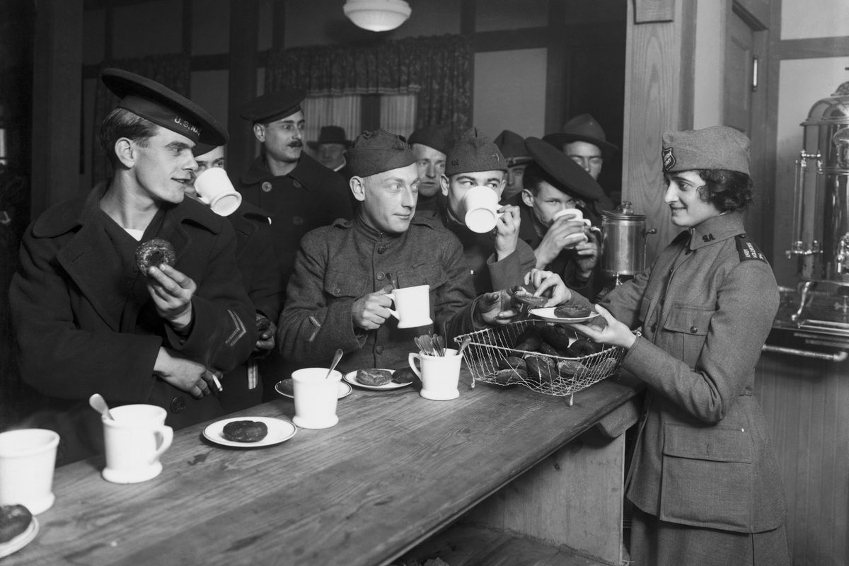 A group of soldiers congregates at a bar to sip coffee and eat doughnuts as a woman working with the Salvation Army distributes the o-shaped confections to the crowd of hungry-looking men.