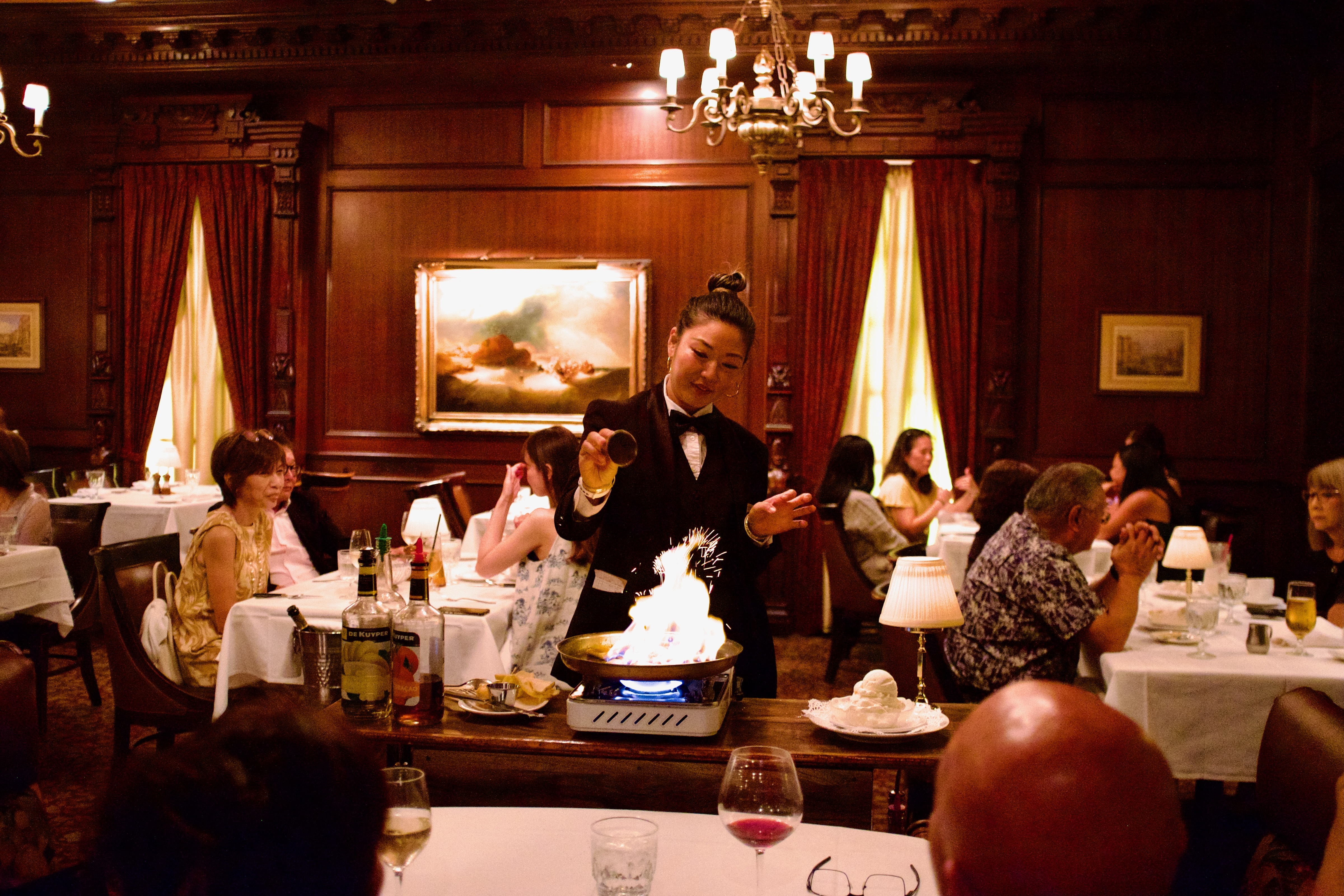 A server stands back as a pan ignites with a large ball of fire, standing in the middle of a midcentury-style wood-lined dining room.