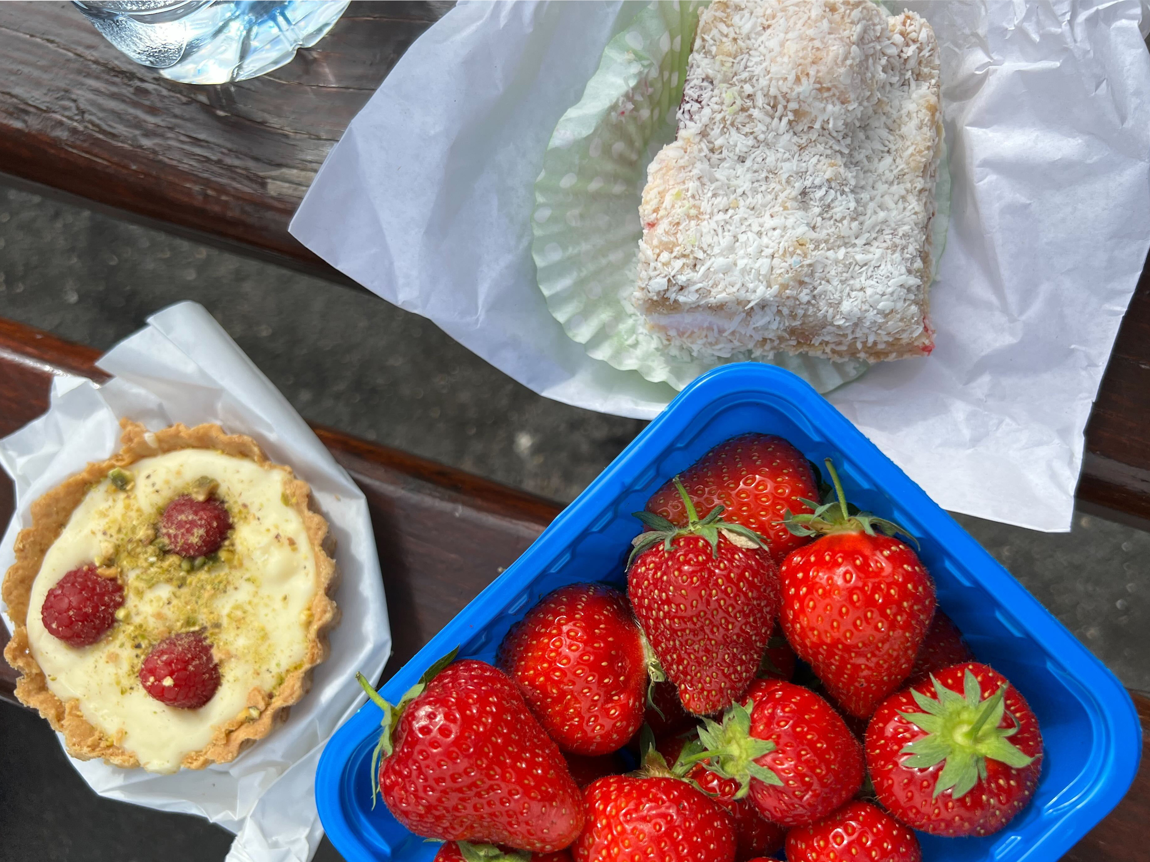 A picnic spread shows an array of coconut-dusted cookies, ultra-ripe strawberries, and a cream tart studded with three raspberries spread out on a picnic blanket.