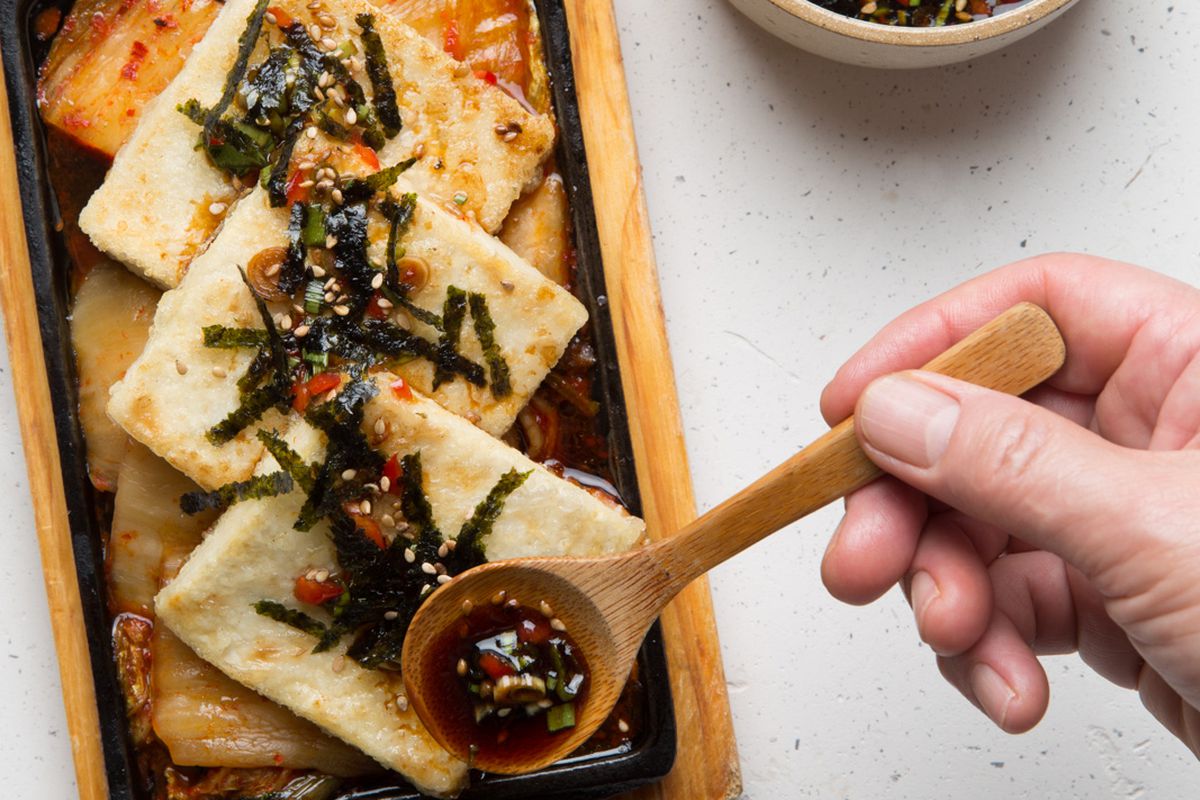Slices of tofu steak with kimchi laid out on a wooden cutting board, with a small bowl of sauce.