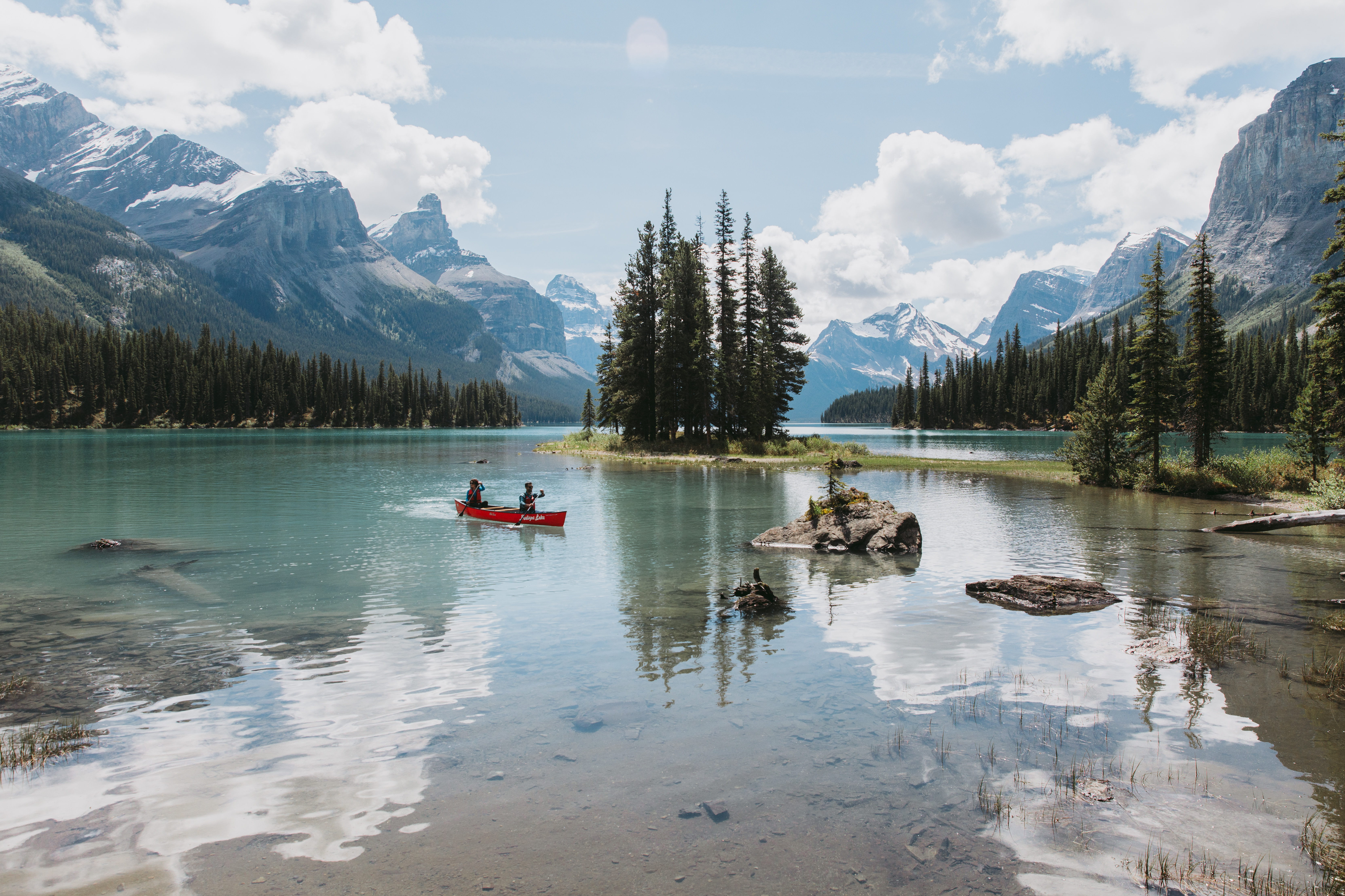 Two people in a small red boat on a mirror-like lake, surrounded by small islands and massive mountains beyond. 